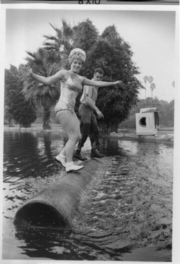 Photo of a woman in a bathing suit, and a lumberjack rolling a log in a pond with an electric dryer on the bank in the background. '